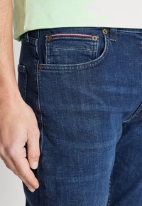 Mörkblå denimjeans med slim fit, som har koppardetaljer, ett röd- och vitt randigt inslag i fickan samt en mjuk textur.