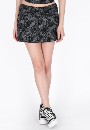 Nő fekete csipkés virágmintás rövid szoknyában és fekete magas szárú sportcipőben fehér fűzőkkel, keresztbe tett lábbal áll.