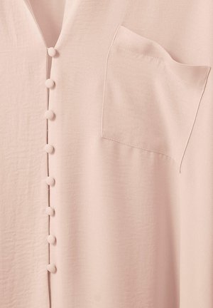 Hellrosa Kleidungsstück mit stoffbezogenen Knöpfen in einer vertikalen Reihe und einer kleinen Brusttasche auf der rechten Seite.