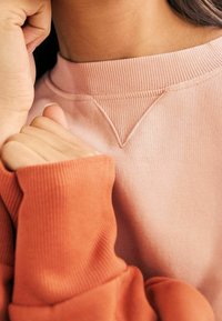 Różowy sweatshirt z żeberkowym dekoltem i teksturowaną łatką. Pomarańczowy rękaw pokazuje bezszwowe dzianie i detale mankietu. Zbliżenie na tkaninę i dłonie.