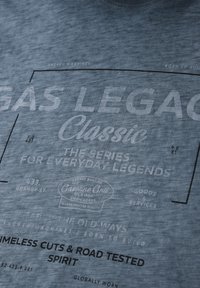 Szara tkanina z wyblakłym białym i czarnym nadrukiem, zawierającym napisy „Gas Legacy Classic”, „Seria dla codziennych legend” oraz „Ponadczasowe kroje i duch testowany na drodze”.