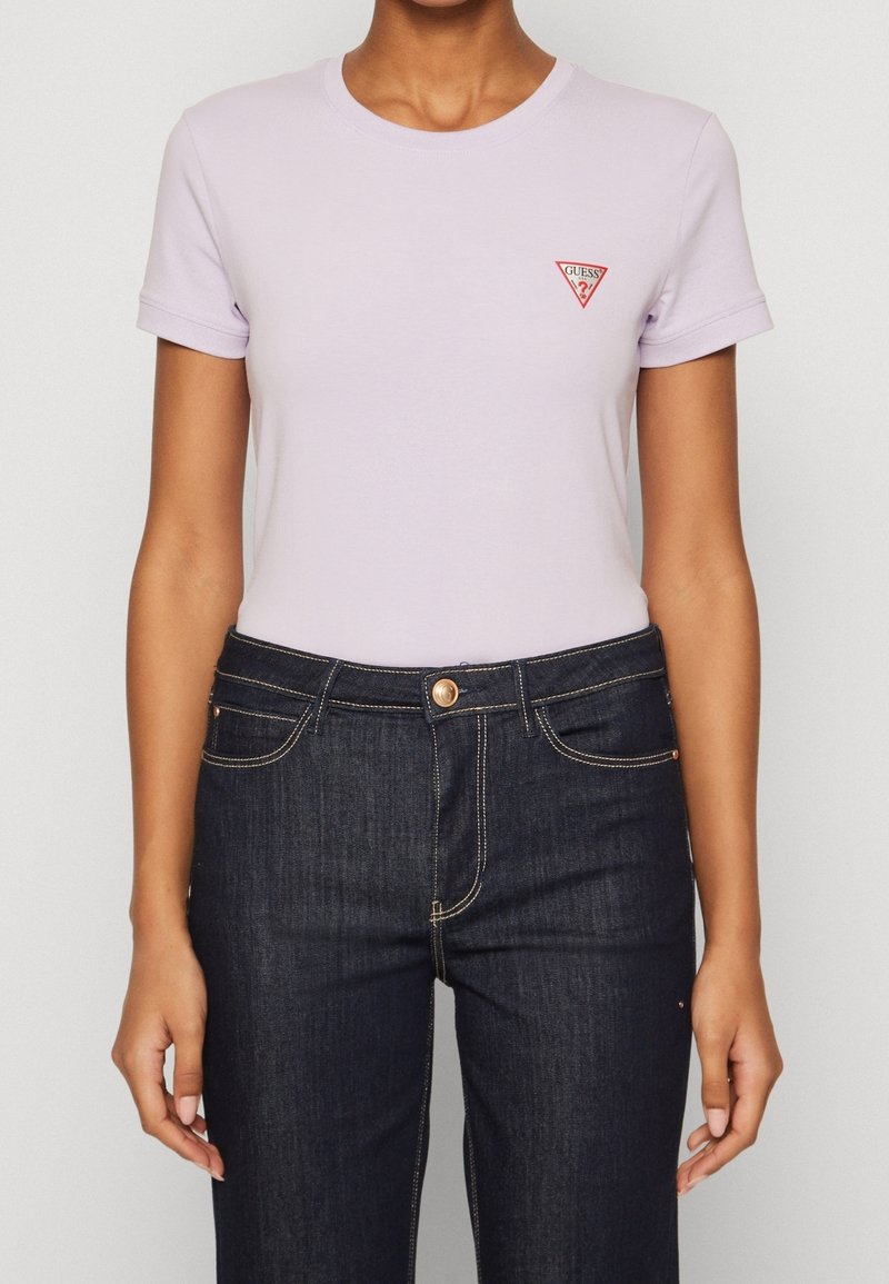 Personne portant un t-shirt à manches courtes violet clair avec un petit logo rouge Guess et un jean taille haute bleu foncé.