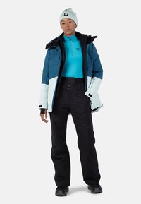 Geïsoleerde skijas in donker teal en lichtblauw met een rits, gecombineerd met zwarte skibroeken en een lichtblauwe muts. Geweven stof.