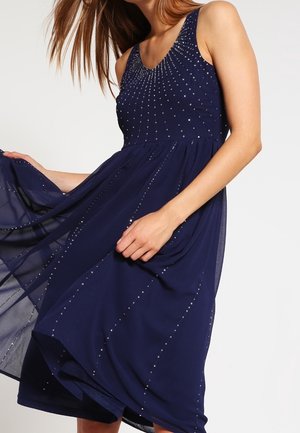 Robe sans manches bleu marine avec un corsage ajusté, des accents de perles scintillantes et une jupe fluide qui oscille doucement. Design élégant avec une texture douce.