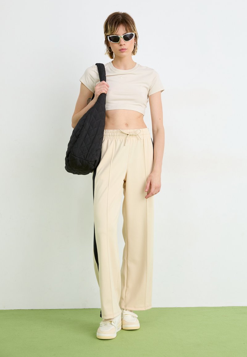 Camiseta recortada beige, pantalones beige sueltos con franjas laterales negras, llevando un bolso de hombro negro acolchado. Zapatillas blancas y gafas de sol de ojo de gato.