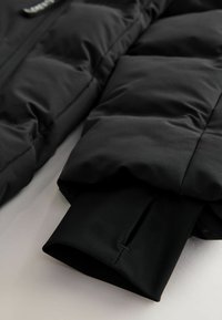 Schwarze Pufferjacke mit glatter Textur, ausgestattet mit einem Reißverschluss vorne und elastischen Ärmelöffnungen für eine enganliegende Passform.