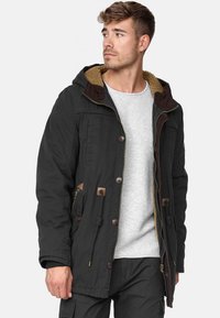 Parka negra con capucha forrada de piel sintética marrón, cierre de cremallera, botones y cordones. Cuenta con bolsillos laterales y acentos en contraste.
