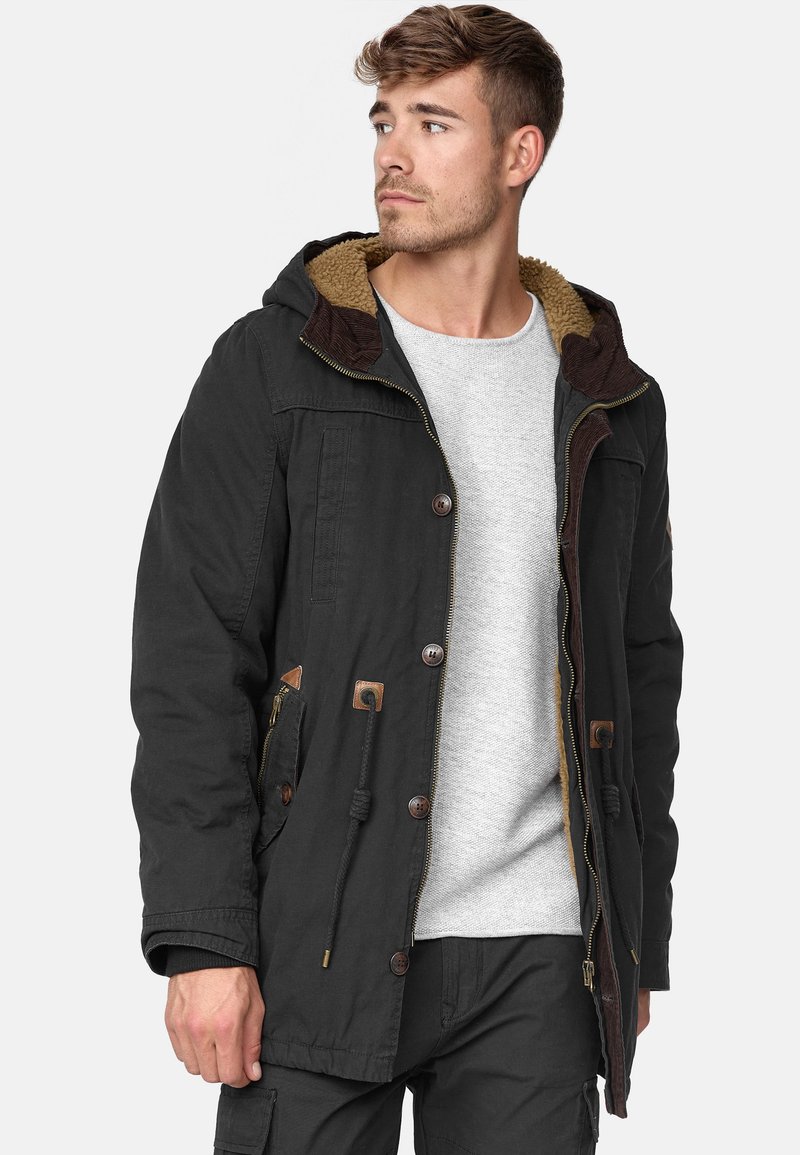 Parka negra con capucha forrada de piel sintética marrón, cierre de cremallera, botones y cordones. Cuenta con bolsillos laterales y acentos en contraste.