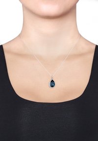 Sølvsmykke med en tåreformet blå ædelsten pendants, omgivet af små klare accenter, båret på en mørk top.