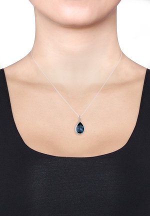 Sølvsmykke med en tåreformet blå ædelsten pendants, omgivet af små klare accenter, båret på en mørk top.