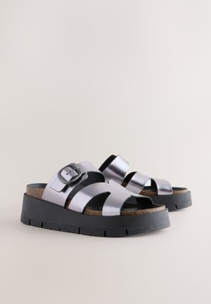 Sandale cu platformă metalice argintii, cu barete încrucișate, detaliu cu cataramă, talpă intermediară din plută și talpă de cauciuc neagră. Textură netedă.