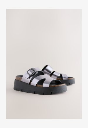 Sidabrinės metalinės platforminės basutės su kryžminėmis dirželiais, sagties detale, kamštinio vidpadžio dalimi ir juodu guminio pado sluoksniu. Lygios tekstūros.