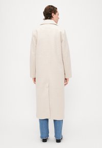 Beige ullblanding lang kåpe med notert krage, rett snitt og baksliss, sammen med lyseblå rette jeans og svart fottøy.