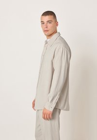 Mladić s kratkom kosom u bež kostim od manšetrnog samta i odgovarajućim hlačama, stoji bočno uz jednostavnu svijetlu pozadinu.