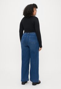Mulher vestindo uma blusa preta justa de mangas longas e jeans de denim azul de perna larga com cintura alta, dois bolsos traseiros e cintura larga.