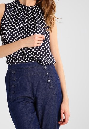 Femme portant un chemisier noir sans manches à pois blancs, rentré dans un pantalon bleu marine taille haute avec des détails de boutons sur le devant.