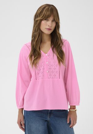 Kobieta z długimi brązowymi włosami, ubrana w różową teksturowaną bluzkę z koronkowymi detalami i niebieskie dżinsy, stojąca na tle jednolitego jasnego tła.