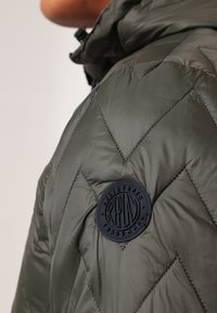 Prošívaná bunda ve tmavě olivové barvě, s prošívaným vzorem, zipem a kulatou černou logovou nášivkou na rameni. Hladká textura.