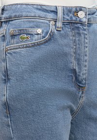 Μπλε τζιν από denim με μπροστινή τσέπη που διαθέτει μικρό κεντητό πράσινο λογότυπο κροκόδειλου και ασημένιο κλείσιμο με κουμπί.