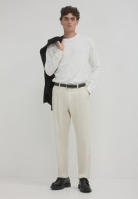 Camicia bianca a maniche lunghe, pantaloni crema, cintura nera e scarpe nere. Il modello tiene un giacca nera sulla spalla, in piedi di fronte a uno sfondo neutro.