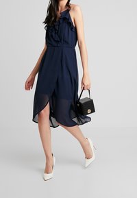 Robe bleu marine avec un col à volants, une sur-jupe en voile transparent et un ourlet asymétrique. Associ ée à un petit sac à main noir texturé et des talons blancs.