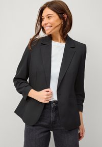 Zwarte blazer met lange mouwen, sluiting met een enkele knoop en opstaande revers. Gecombineerd met een lichtgrijze shirt en hoge taille donkerblauwe jeans.