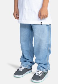 Světle modré džíny s volným střihem, s viditelným šitím a černou nášivkou. Oblečeno s šedými teniskami a bílými tkaničkami.