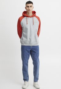 Sudadera con capucha gris y roja, con un bolsillo tipo canguro, puños acanalados y cordón. Combinada con jeans azules y zapatillas blancas.