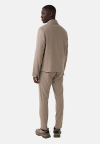 Mand iført beige skræddersyetjakke og bukser, parret med kraftige beige og sorte sneakers, står sidelæns foran en hvid baggrund.