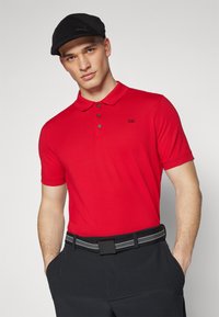 Crvena polo majica od pamuka s ovratnikom, kratkim rukavima i tri gumba. Na lijevoj strani grudi ima crni logotip s brendom.