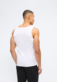 Άνδρας που στέκεται πλάτη, φορώντας άσπρο αμάνικο τοπ και μαύρο παντελόνι, μπροστά σε απλό λευκό φόντο.