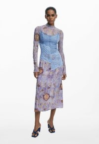 Vestido de mezclilla y encaje en azul y lavanda con patrones florales; diseño de mangas largas y media longitud. Las sandalias cuentan con tiras azules contrastantes.