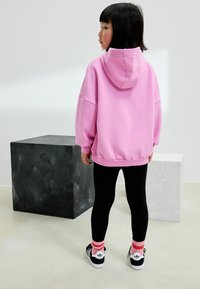 Różowa bluza z dużym kapturem, luźny krój, noszona na czarnych legginsach, w połączeniu z białymi sneakersami i różowymi skarpetkami w paski. W tle dwa geometryczne bloki.