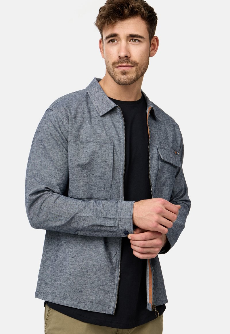 Grau gewebte Hemdjacke mit Kragen, langen Ärmeln, einer Brusttasche und orangefarbener Reißverschlussdetails. Der Stoff hat eine strukturierte Oberfläche.
