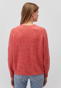 Korallefarbener Strickpullover mit einem strukturierten Zickzackmuster auf der Rückseite, langen Ärmeln und einem runden Ausschnitt, kombiniert mit hellblauen Jeans.