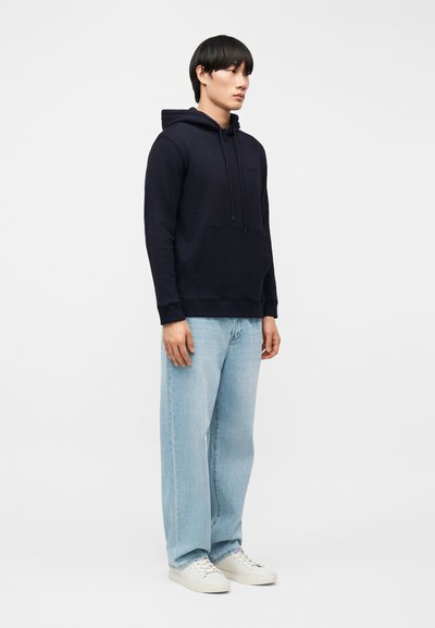 Άνδρας στέκεται φορώντας μαύρο φούτερ με κουκούλα, ανοιχτό μπλε τζιν φαρδιάς γραμμής και λευκά αθλητικά παπούτσια μπροστά σε απλό λευκό φόντο.