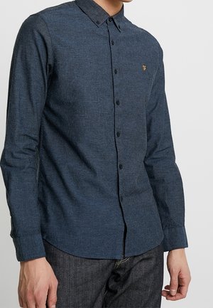 Mann trägt ein eng anliegendes, dunkelblaues Hemd mit langen Ärmeln und einem kleinen, auf der Brust gestickten Logo.