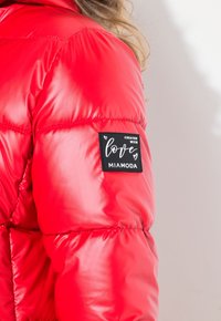 Chaqueta de plumas roja con un acabado brillante, que presenta un parche cuadrado etiquetado "Creado con amor" y "MIAMODA" en la manga izquierda.