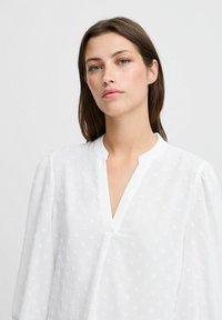 Valge blousee V-kaelusega, kerge kangas tekstuursete täppidega ja puhvvarrukatega. Lihtne ja elegantne disain.