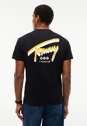 Homme vu de dos portant un t-shirt noir avec un grand logo Tommy NYC jaune et blanc et un texte au dos, associé à un jean bleu.
