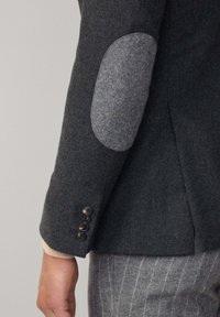 Blazer in lana grigia con toppe in feltro grigio sui gomiti, dettaglio a cinque bottoni sui polsini e una forma sartoriale con superficie testurizzata.