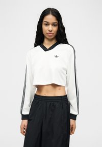 Mlada ženska z dolgimi temnimi lasmi, oblečena v belo obrezano Adidas trenirko s črnimi črtami in široke črne hlače, stoji na belem ozadju.