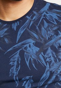T-shirt marine avec un imprimé botanique en bleu clair. Le design présente des feuilles détaillées et un col rond. Le tissu semble doux et texturé.