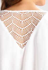 Vêtement blanc avec un dos délicat en dentelle présentant une incrustation triangulaire. La dentelle a un motif géométrique et une finition texturée.