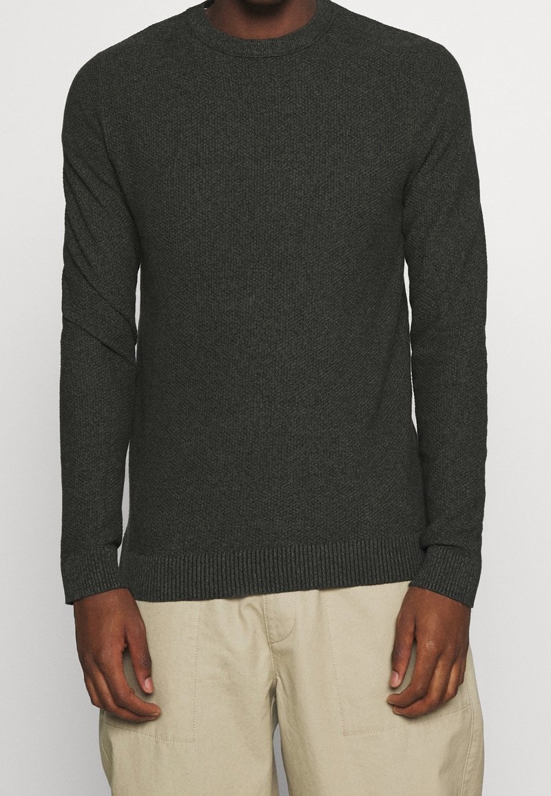 Pull en maille vert foncé avec un col ras du cou, des manches longues et des bords côtelés au niveau des poignets et de l'ourlet. Le motif texturé ajoute un intérêt visuel.