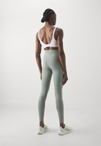 Bra sportivo bianco con chiusura posteriore regolabile, abbinato a leggings verde chiaro a vita alta. Modello con scarpe da ginnastica bianche.
