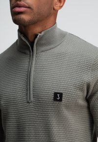 Grijze ritstrui met een textuurpatroon, hoge kraag en zwart logo-opdruk op de linkervoorkant. Materiaal lijkt zacht en gebreid.