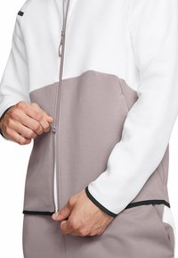 Reißverschluss-Sportjacke in Weiß und Hellgrau. Merkmale eine glatte Textur, ein Farbblockdesign und schwarze Akzente am Saum und an den Bündchen.