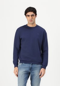 Felpa in cotone blu navy con polsini e colletto a costine, caratterizzata da un logo impresso discreto sul petto. Abbinata a jeans azzurri chiari.