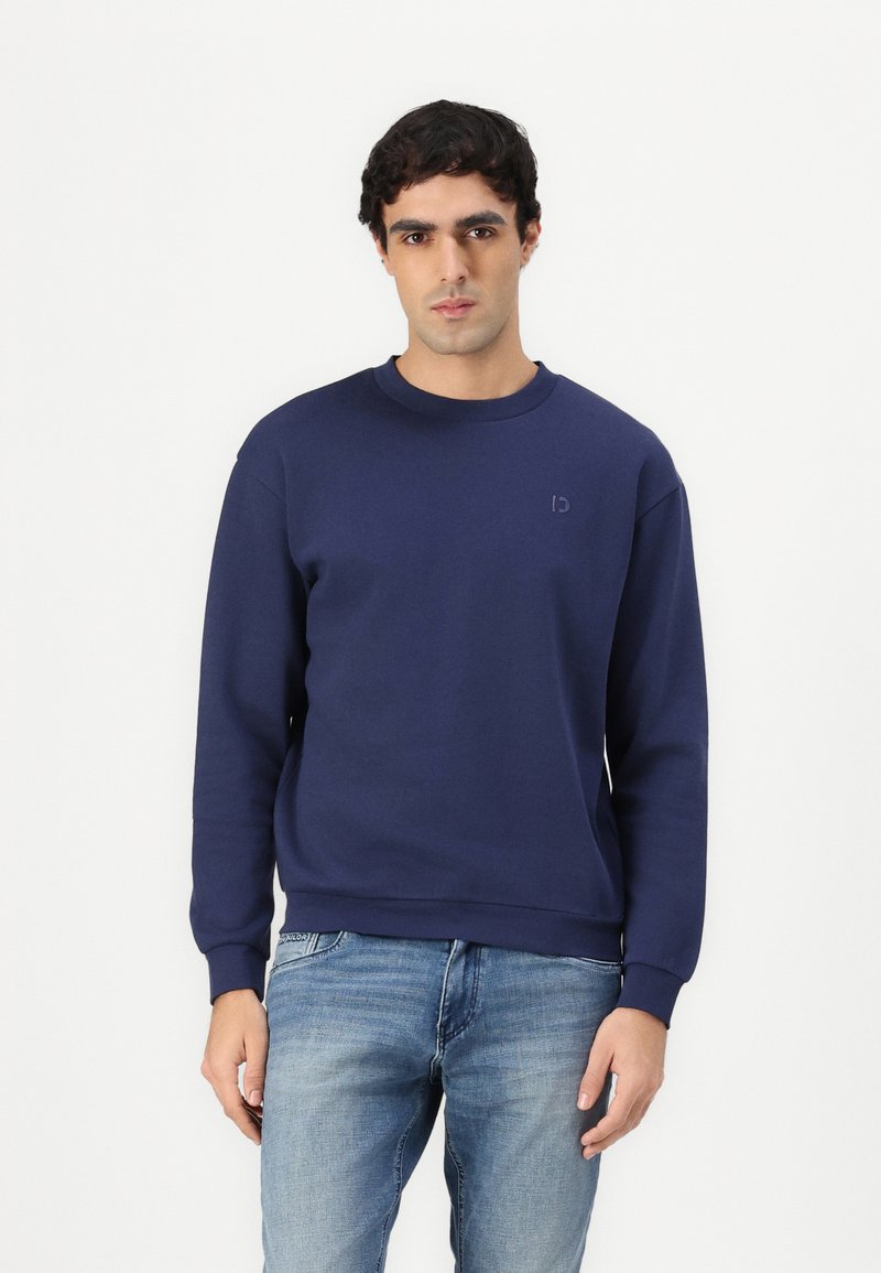 Felpa in cotone blu navy con polsini e colletto a costine, caratterizzata da un logo impresso discreto sul petto. Abbinata a jeans azzurri chiari.